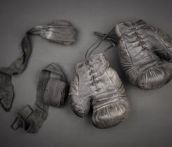 Worn black leather boxing gloves with visible creases and laces, laid flat beside faded hand wraps on a dark gray background.