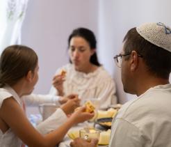 A family sits at a table eating a meal, with a man wearing a kippah and a child sharing food.