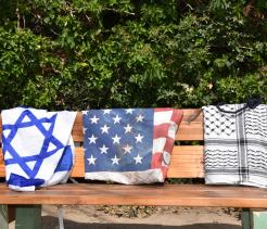 Three cloths draped over a bench: the Israeli flag, the U.S. flag, and a black-and-white keffiyeh.