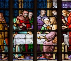 Colorful stained-glass window depicting a medieval scene of several robed figures gathered around a table, with expressive faces and ornate architectural details in the background.