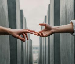 Two outstretched hands reaching toward each other between tall concrete memorial slabs, symbolizing connection, memory, and loss.