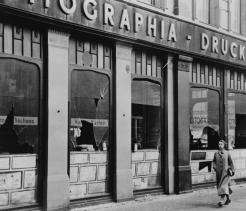 Black-and-white photograph of a storefront with shattered windows and debris on the sidewalk. Large lettering above the windows includes the word “Druckerei,” and a woman in a long coat walks past the damaged building.