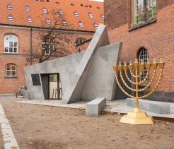 Large outdoor menorah sculpture standing in a courtyard beside a modern concrete memorial structure. The setting is surrounded by historic red brick buildings with arched windows, creating a contrast between contemporary memorial design and older architecture. Fallen leaves on the ground suggest an autumn setting.
