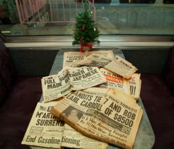 Stack of vintage newspapers with World War II headlines about Japan’s surrender spread across a train table, with a small decorated Christmas tree placed by the window behind them.