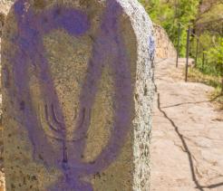 Weathered stone marker with a carved menorah symbol, set along a natural stone pathway bordered by metal railings and surrounded by greenery.