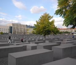 Rows of large gray concrete blocks of varying heights form a grid-like pattern at the Holocaust Memorial in Berlin, with a green tree rising among the structures and city buildings visible in the background.