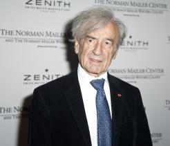 Portrait of an elderly man with gray hair wearing a dark suit and blue tie, standing in front of a step-and-repeat backdrop with event and sponsor logos.