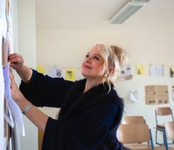 A smiling woman in a classroom pins papers to a bulletin board, with empty desks and student work displayed on the wall behind her.