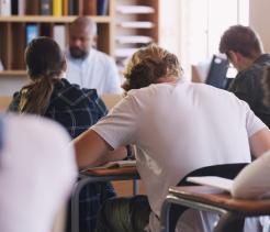 Students seated at desks in a classroom, viewed from behind, writing in notebooks while a teacher stands at the front near a bookshelf. The scene shows a focused learning environment with several students taking notes during a lesson.