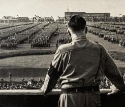 Black-and-white historical photograph taken from behind a uniformed man standing on a raised platform or balcony, looking out over a massive, tightly organized crowd assembled in long rectangular formations. The man wears a military-style uniform with a dark armband. In the distance are large buildings and multiple flags on tall poles. The scene suggests a large political rally or military assembly in an urban setting.
