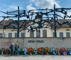 Color photograph of a memorial site with a large abstract metal sculpture of human figures suspended above a concrete wall engraved with the years “1933–1945.” Numerous wreaths with colorful ribbons are arranged along the base of the wall. Two people stand in front of the memorial, appearing to read or pay their respects. A building with a sloped roof and multiple windows stands behind the monument under a partly cloudy sky.