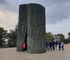 A war memorial featuring bronze statues of soldiers emerging from a broken brick wall, depicted in motion as if advancing during battle. The soldiers wear military uniforms and helmets, and one has a red-and-white armband. Numerous wreaths and bouquets with red and white ribbons are arranged in front of the monument. The sculpture is set in a paved plaza, with modern glass buildings and tall green columns in the background.