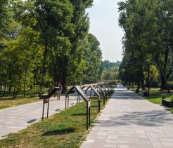 A wide paved walkway in a leafy park lined with tall trees, informational signboards on metal stands, and modern black lampposts, with a few people walking and sitting along the path on a sunny day.