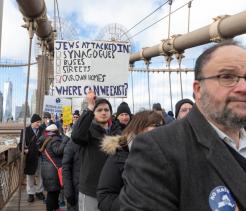 A group of people participate in a protest march across a large suspension bridge. In the foreground, a middle-aged man wearing glasses and a dark coat has a “No Hate” button pinned to his jacket. Behind him, a young man holds a sign that reads, “Jews attacked in: synagogues, buses, streets, our own homes. Where can we exist?” Other participants wear winter clothing and hold additional signs. City buildings and bridge cables are visible in the background.