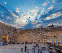 Wide view of the Western Wall plaza in Jerusalem at sunset, with people gathered in prayer near the stone wall, rows of white chairs and folded umbrellas in the foreground, and birds flying across a dramatic sky with sun rays breaking through the clouds.