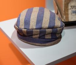 A small blue-and-gray striped fabric cap displayed on a white museum pedestal against an orange background, resembling a prisoner’s uniform hat, with a worn paper label visible behind it.