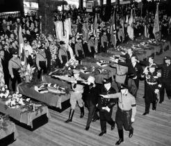 Black-and-white historical photograph of Nazi officials and uniformed soldiers performing the Nazi salute during a formal ceremony, with multiple flag-draped coffins arranged in rows inside a large hall and a crowd observing in the background.