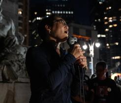 A person holding a microphone speaks at a nighttime outdoor event in a city. Streetlights and illuminated buildings are visible behind them, and a few people stand nearby listening.