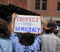 A person in a crowd holds a handmade cardboard sign above their head that reads “PROTECT OUR DEMOCRACY” in red and blue letters with stars. The crowd is gathered outdoors in front of brick buildings, suggesting a public protest or demonstration.