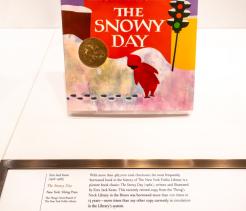 A children’s picture book titled The Snowy Day by Ezra Jack Keats is displayed upright in a museum-style case. The book cover shows a small child in a red snowsuit standing in a snowy city scene, with bold, colorful shapes and a gold award medallion on the front. Below the book, a placard provides explanatory text about its historical and cultural significance.