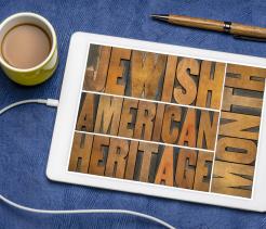 A tablet lies on a blue fabric surface displaying the words “Jewish American Heritage” arranged in bold wooden letterpress blocks. Nearby are a yellow mug filled with coffee, a wooden pen, and a white charging cable, creating a casual desk or tabletop setting that suggests reflection, learning, or celebration of Jewish American heritage.