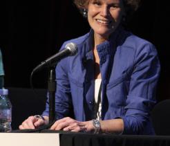 Author Judy Blume sits at a panel table speaking into a microphone, smiling warmly toward the audience. She wears a blue jacket over a light top, with short curly hair, and a name placard reading “Judy Blume” is visible in front of her against a dark stage background.