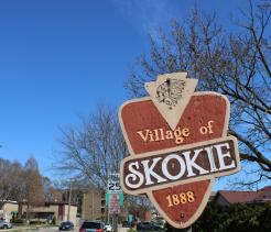 Wooden roadside sign reading “Village of Skokie, 1888” with a Native American profile emblem at the top, set against a clear blue sky. Bare tree branches, nearby houses, parked cars, and street signs are visible in the background, indicating a suburban neighborhood.