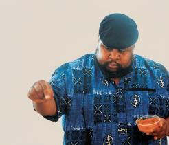 A man wearing a blue patterned shirt and a dark cap sprinkles seasoning from one hand while holding a small bowl of sauce in the other, focused on the action against a plain light background.
