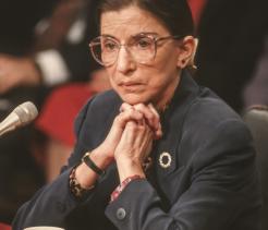 A woman wearing glasses sits at a hearing table with her hands clasped near a microphone, listening attentively.