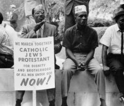 Black-and-white photograph of several men seated outdoors during a civil rights era demonstration. One man holds a sign reading, “We march together Catholic Jews Protestant for dignity and brotherhood of all men under God NOW!” while others sit beside him wearing hats and dress shirts.