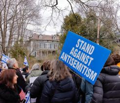 A crowd of people gathers outdoors in a residential neighborhood during winter, many wearing coats. One person holds a large blue sign reading “STAND AGAINST ANTISEMITISM.” Several attendees hold small Israeli flags, and leafless trees and houses are visible in the background, suggesting a peaceful demonstration or rally.
