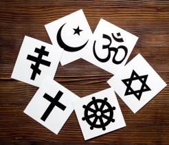 Overhead view of paper cards arranged in a circle on a wooden surface, each displaying a different religious symbol, including a cross, crescent and star, Om, Star of David, and Dharma wheel.