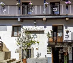 A sunlit street scene shows a traditional whitewashed building in the Jewish quarter of Hervás, Spain. The façade features balconies decorated with flower boxes and metal railings displaying Star of David symbols. Below, a small plaza area includes a stone fountain, a menorah sculpture, and Jewish symbols mounted on the wall. An open doorway leads into a shop, reinforcing the sense of a preserved historic neighborhood that reflects Jewish cultural heritage and daily life.