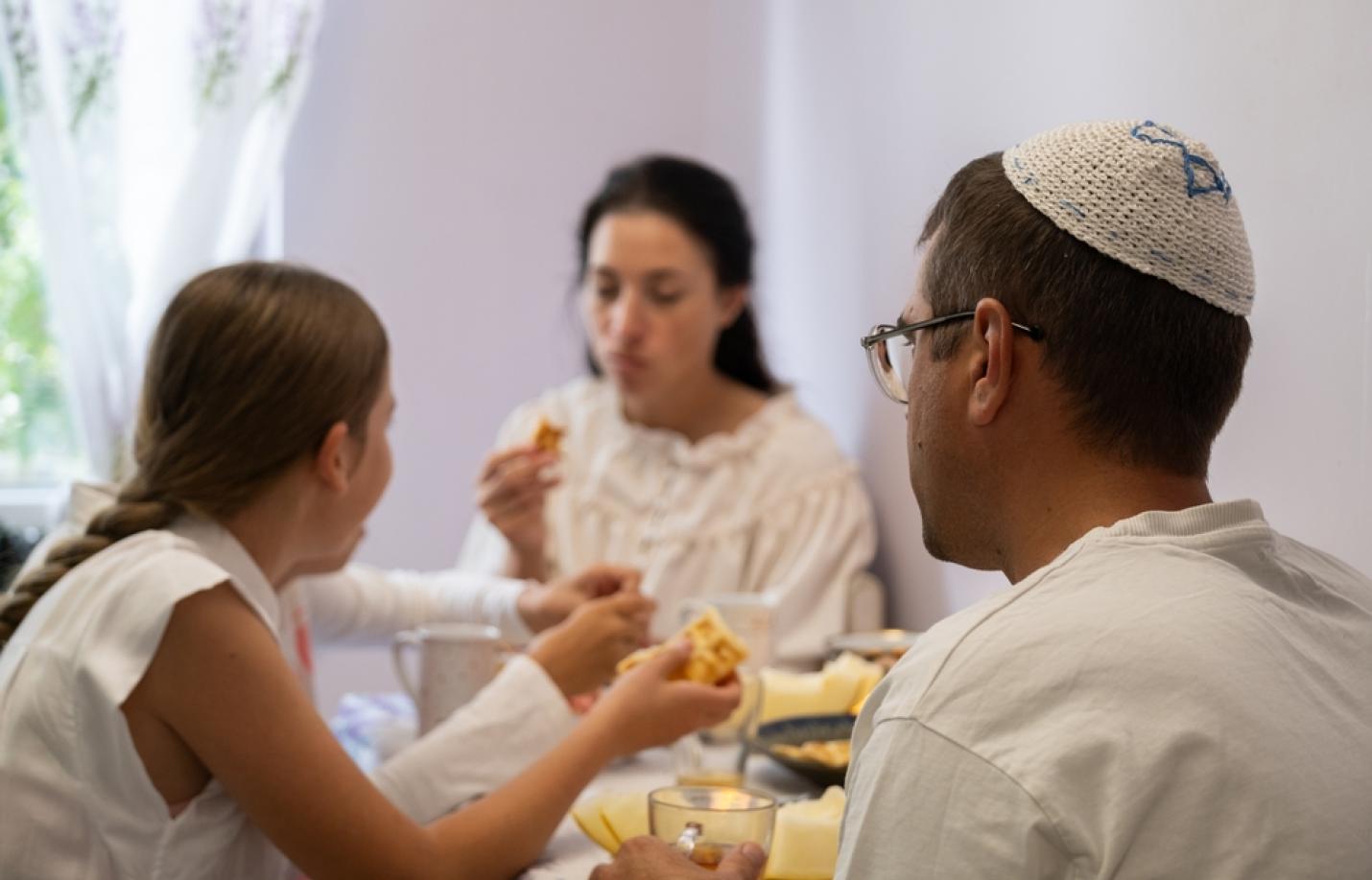A family sits at a table eating a meal, with a man wearing a kippah and a child sharing food.