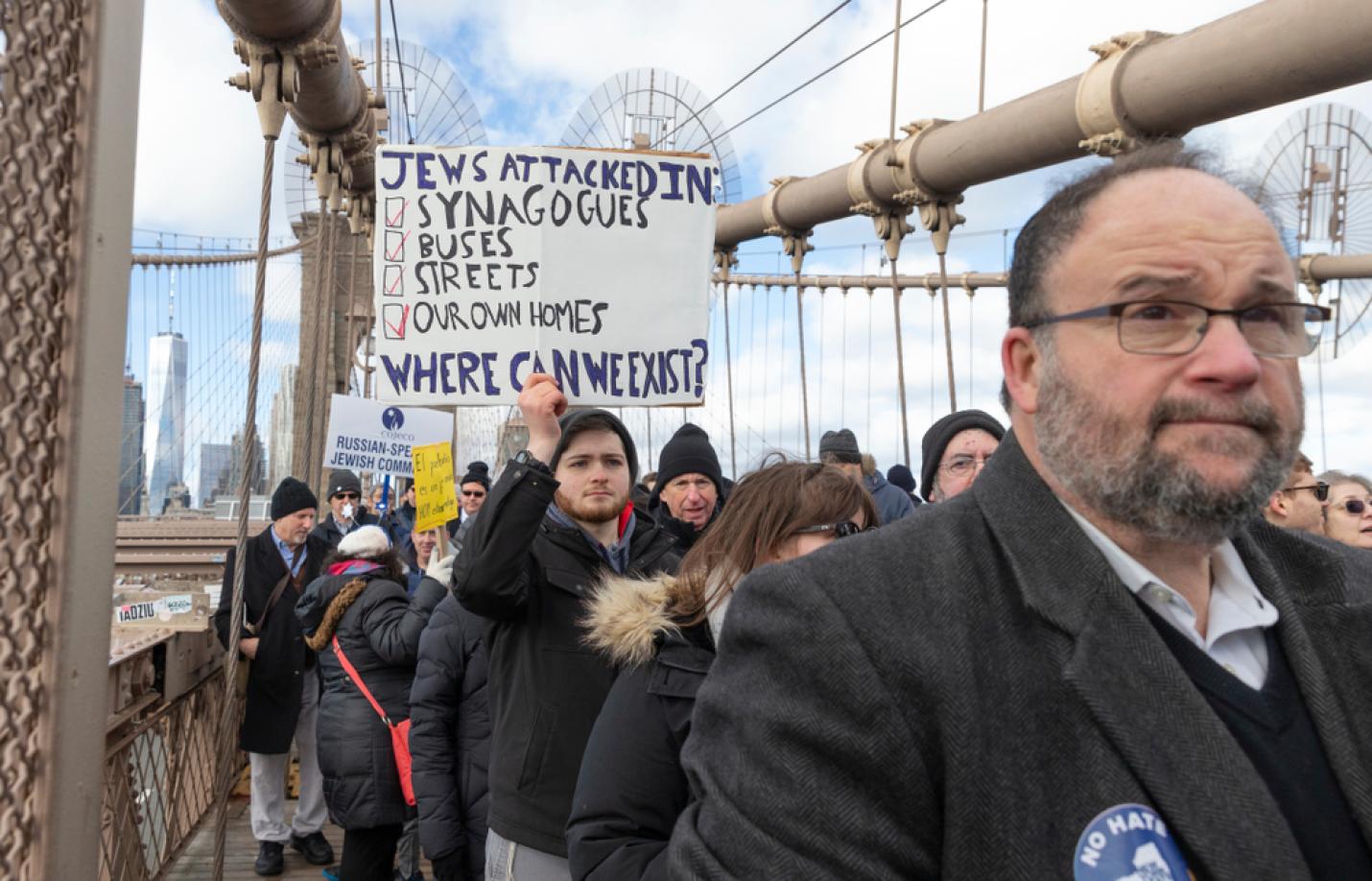 A group of people participate in a protest march across a large suspension bridge. In the foreground, a middle-aged man wearing glasses and a dark coat has a “No Hate” button pinned to his jacket. Behind him, a young man holds a sign that reads, “Jews attacked in: synagogues, buses, streets, our own homes. Where can we exist?” Other participants wear winter clothing and hold additional signs. City buildings and bridge cables are visible in the background.