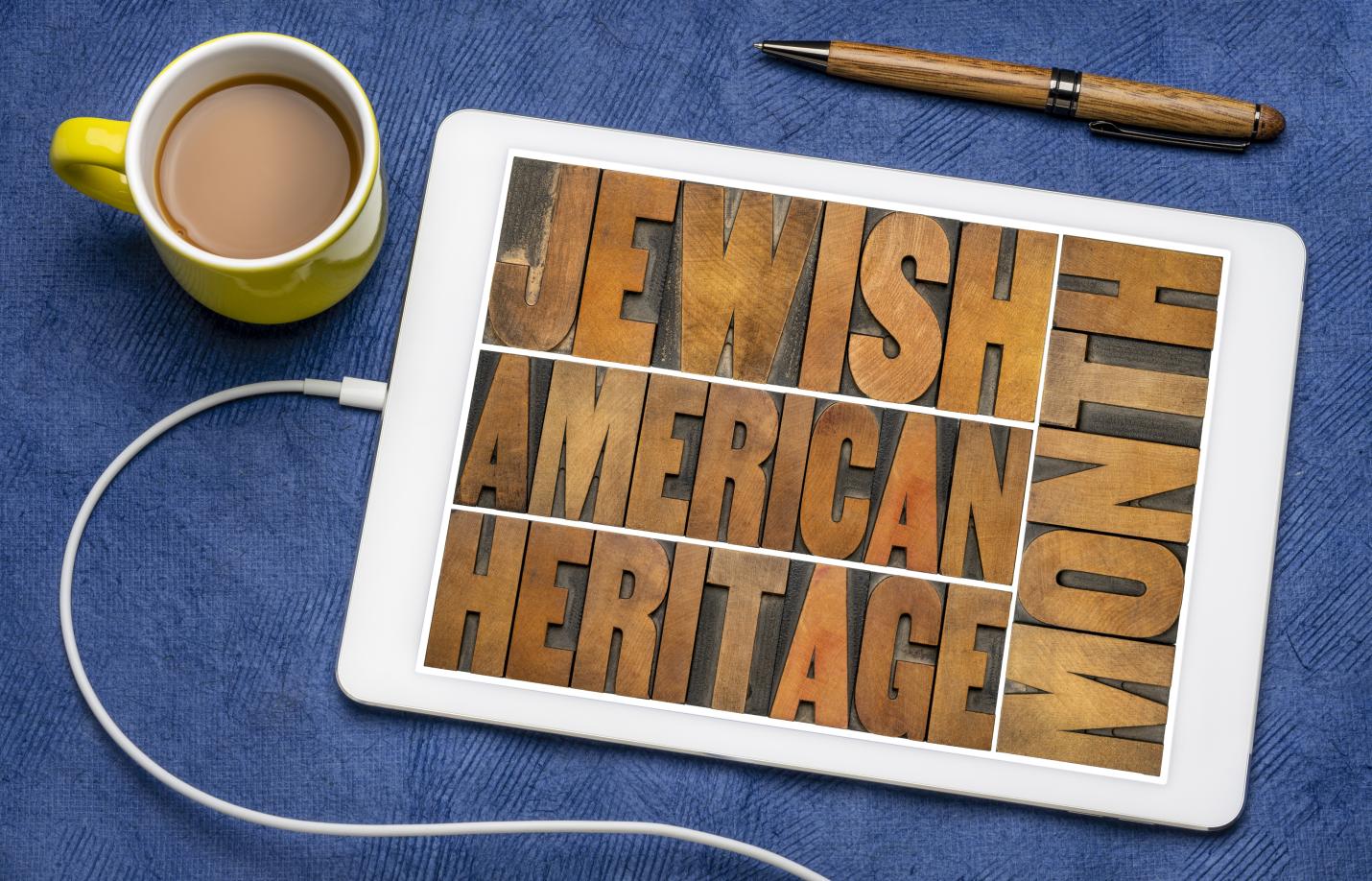 A tablet lies on a blue fabric surface displaying the words “Jewish American Heritage” arranged in bold wooden letterpress blocks. Nearby are a yellow mug filled with coffee, a wooden pen, and a white charging cable, creating a casual desk or tabletop setting that suggests reflection, learning, or celebration of Jewish American heritage.