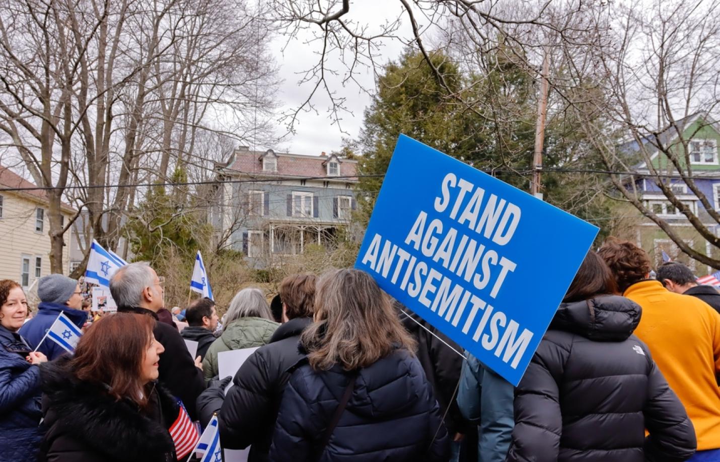 A crowd of people gathers outdoors in a residential neighborhood during winter, many wearing coats. One person holds a large blue sign reading “STAND AGAINST ANTISEMITISM.” Several attendees hold small Israeli flags, and leafless trees and houses are visible in the background, suggesting a peaceful demonstration or rally.